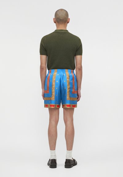 Άνδρας που φοράει πράσινο κοντομάνικο πουκάμισο, φωτεινά μπλε σορτς με κόκκινες και χρυσές λεπτομέρειες, άσπρες κάλτσες και μαύρα παπούτσια, στέκεται γυρισμένος με την πλάτη.