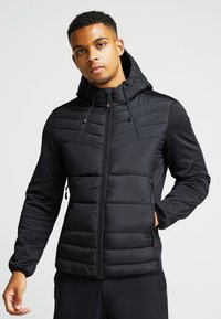 Giacca isolante nera con zip frontale, cappuccio e pannelli texture. Presenta polsini elastici e tasche laterali, realizzata con materiali morbidi.