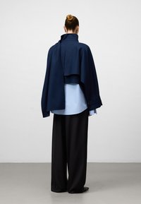 Un cappotto esterno blu navy si sovrappone a una camicia azzurro chiaro, abbinato a pantaloni neri larghi. Texture liscia, design minimalista con dettagli sottili.