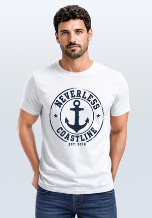 Mann mit dunklem lockigem Haar und Bart, der ein weißes T-Shirt mit marineblauem Anker-Logo und Text "Nevertheless Coastline Est. 2016" trägt, steht mit den Händen hinter dem Rücken.