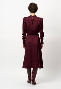 Bordeaux satijnen jurk met lange mouwen, een keyhole achterkant, een getailleerde taille en een vloeiende rok. Gepaard met glanzende, kniehoge bordeaux laarzen.
