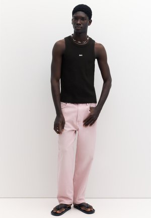Jeune homme debout portant un débardeur noir, un pantalon rose, un collier de perles et des sandales noires sur un fond blanc uni.