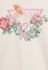 Valkoinen t-paita, jossa on vaaleanpunainen kimaltava kolmionmuotoinen logo kukkakuviolla, johon kuuluu erilaisia värikkäitä kukkia ja vihreitä lehtiä.