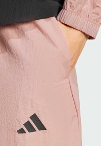 Light pink sports pants with a crinkled texture, featuring a black Adidas logo and a partially visible black top with gathered cuffs.