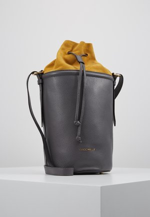 Ciemnoszara skórzana torba typu bucket z musztardowożółtym zamszowym zamknięciem na ściągacz i cienkim, regulowanym paskiem na ramię na białym tle.