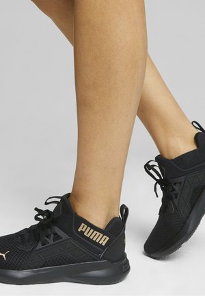 Personne portant des chaussures de sport noires Puma avec des détails en mesh et géométriques, montrée de profil et de dos sur un fond uni.