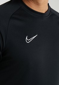 Muž v černém tričku s kulatým výstřihem a bílým logem Nike swoosh na hrudi, bez dalších viditelných detailů.