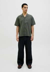 Chemise à manches courtes, couleur olive, avec des boutons, associée à un jean en denim foncé à jambes larges et des chaussures brunes. Design simple et moderne.