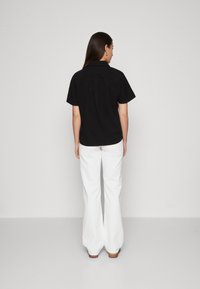 Camicetta nera a maniche corte con colletto, caratterizzata da una plica sul retro, abbinata a pantaloni bianchi a gamba larga e calzature marroni. Design semplice e pulito.