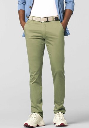 Mann trägt hellgrüne, schmal geschnittene Hose, weißen Gürtel, weiße Sneakers, weißes T-Shirt und hellblaues, geknöpftes Hemd mit Händen in den Taschen.