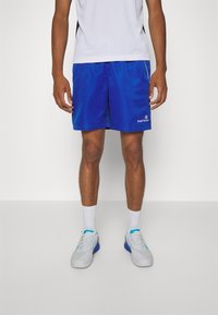 Man som bär blå Sergio Tacchini sportshorts, vita strumpor och vitblå sneakers står mot en enfärgad bakgrund.