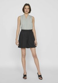 Hellgraues ärmelloses Oberteil mit umgeschlagenem Kragen, kombiniert mit hochtaillierten, lockeren schwarzen Shorts und schwarzen Riemchen-Heels, vor einem neutralen Hintergrund.