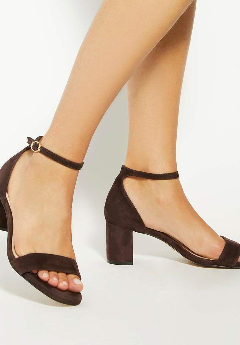Brown suede high-heeled sandals with a block heel, open toe, ankle strap featuring a circular gold accent, and smooth texture.