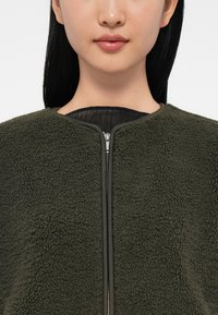 Woman wearing a dark green textured fleece jacket with a front zipper and black inner shirt visible at the collar.