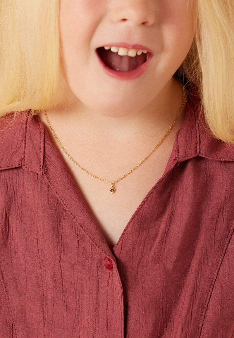Gouden ketting met een kleine hanger, gedragen over een gekreukt bordeauxrood shirt met een v-hals en een rode knoopdetail.