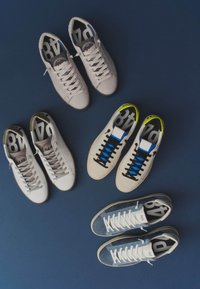 Quatre paires de baskets décontractées à lacets en blanc, écru, crème avec lacets noirs et bleu, disposées sur un fond bleu foncé.
