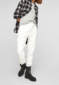 Black and white checkered flannel shirt over a gray t-shirt; white textured pants with drawstring; black ankle boots.