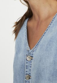 Nainen, joka käyttää vaaleansinistä farkkutoppia, jossa on metalliset napit, joissa lukee "cream denim", ja näkyvissä on kaulan ja olkapään alue sekä osa hiuksista.