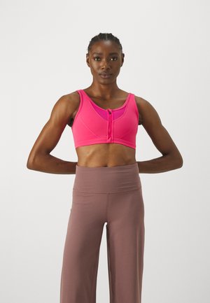 Rosa Sport-BH mit einem Frontreißverschluss, breiten Trägern und unterstützendem Sitz. Getragen mit hochgeschnittenen, hellbraunen Trainingshosen. Glatte Stofftextur.