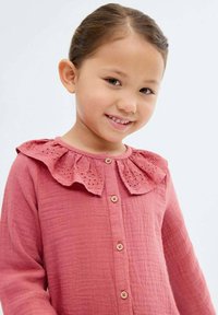 Hymyilevä nuori tyttö, jolla on pitkät hiukset, pukeutuneena vaaleanpunaiseen paitaan, jossa on röyhelökaulus ja puuknopf, vaaleaa taustaa vasten.