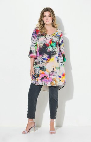 Mehrfarbige Blumen-Tunika mit V-Ausschnitt und dreiviertellangen Glockenärmeln, kombiniert mit dunklen Jeans mit Knopfakzenten an den Knöcheln.