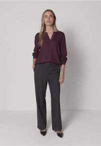 Bordeauxrotes Langarmshirt mit V-Ausschnitt, kombiniert mit hoch taillierter Hose in dunkelgrau. Das Model trägt schwarze spitze Schuhe. Neutraler Hintergrund.
