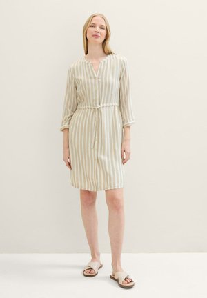 Femme portant une robe à boutons beige et blanche à rayures verticales avec une taille à cordon et des sandales crème à enfiler, se tenant devant un mur uni.