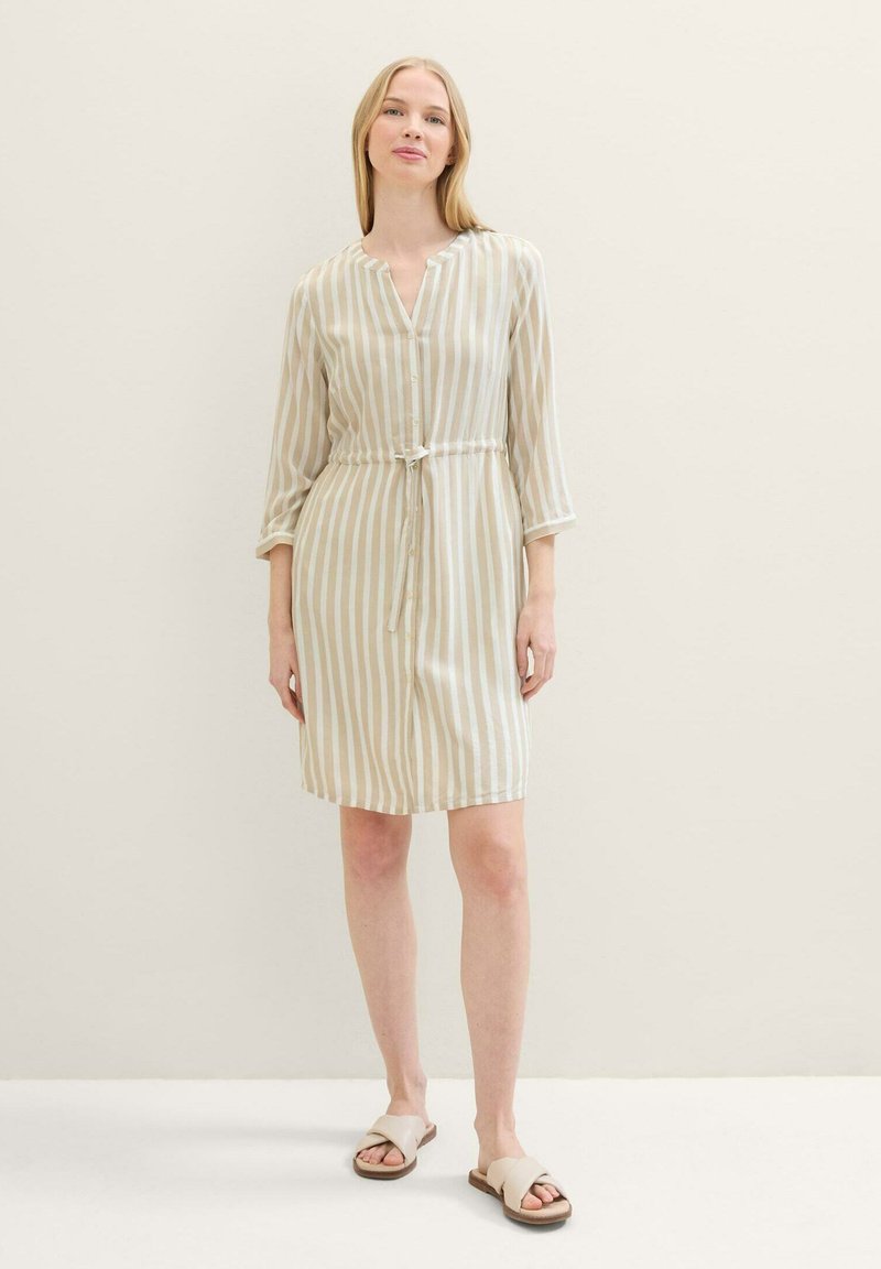 Femme portant une robe à boutons beige et blanche à rayures verticales avec une taille à cordon et des sandales crème à enfiler, se tenant devant un mur uni.
