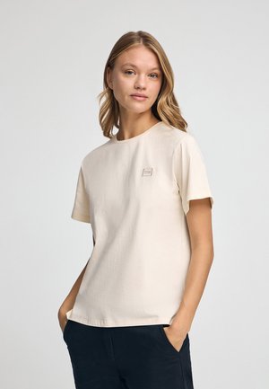 Camiseta de cuello redondo color beige claro, hecha de una tela suave. Cuenta con mangas cortas y un pequeño parche de logo cuadrado en el pecho. Diseño sencillo y minimalista.