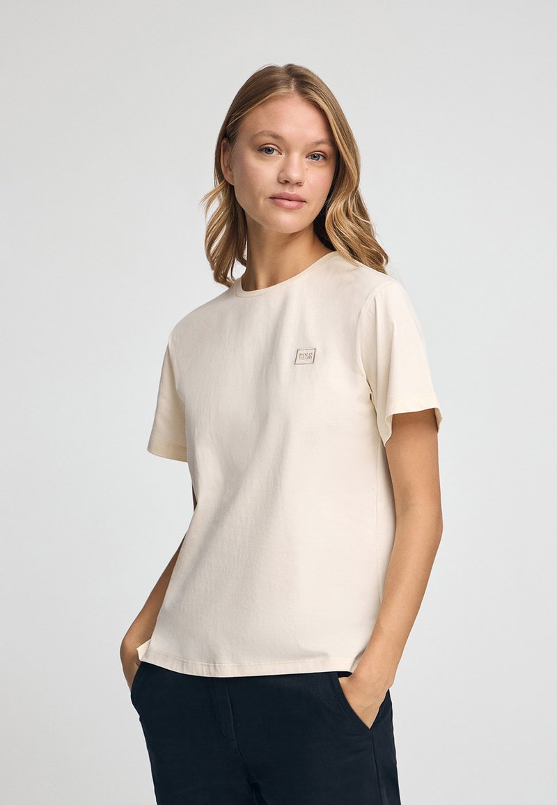 Camiseta de cuello redondo color beige claro, hecha de una tela suave. Cuenta con mangas cortas y un pequeño parche de logo cuadrado en el pecho. Diseño sencillo y minimalista.