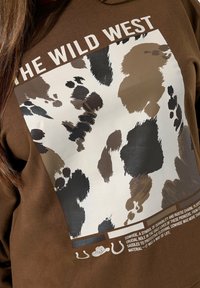 Brązowy sweatshirt z dużym, abstrakcyjnym nadrukiem z krowiej skóry w kolorze beżowym, czarnym i różnych odcieniach brązu, z napisem "DZIKI ZACHÓD" powyżej.