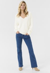Cremfarbener V-Ausschnitt-Pullover mit langen Ärmeln, kombiniert mit blauen Schlagjeans und beigen spitz zulaufenden Schuhen, stehend vor einem schlichten weißen Hintergrund.