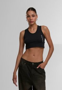 Black cropped tank top made of smooth fabric with a round neckline and subtle side logo detail. Paired with dark, loose-fit jeans.