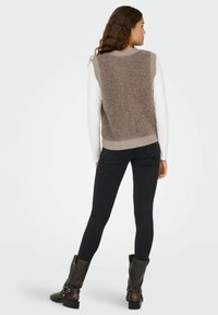 Braun strukturierter ärmelloser Pullover über einem weißen Langarmshirt, kombiniert mit schwarzen Skinny-Jeans und kniehohen schwarzen Stiefeln.