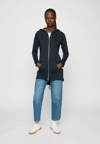 Hoodie zippé bleu marine avec une capuche à cordon, des poches avant et une silhouette plus longue, associé à un jean bleu clair et des baskets blanches.