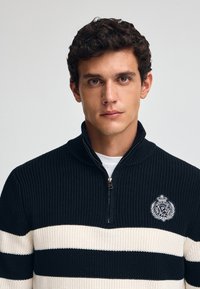 Svart och krämrandig stickad tröja med dragkedja vid kragen och en broderad logotyp på bröstet. Har en texturerad, ribbad materialstruktur.