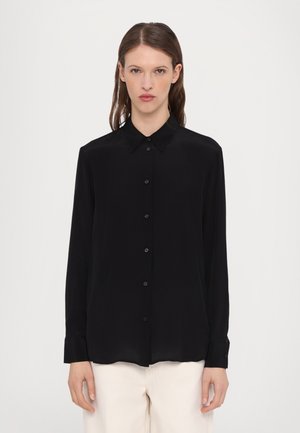 Camicia nera a maniche lunghe con bottoni, tessuto morbido, colletto classico, polsini a singolo bottone, con orlo dritto. Indossata con pantaloni di colore chiaro.