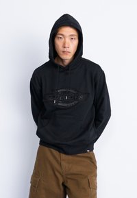 Μαύρη φούτερ με μπροστινή τσέπη, featuring ένα υφασμάτινο λογότυπο και ρυθμιζόμενα κορδόνια. Ο ιστός φαίνεται απαλός και άνετος.