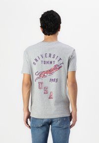 Szürke pamut póló, amelyen egy ugró tigris grafika látható narancssárga és lila színekben, a "University Tommy USA 1985" felirattal hasonló színekben.