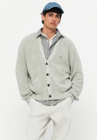 Light gray knitted cardigan with a V-neck, black buttons, and front pockets, layered over a blue and white striped shirt.