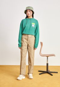 Camicia verde a maniche lunghe con stampa grafica, pantaloni beige a gamba dritta e cappello a secchiello con motivo. Le sneakers completano l'outfit.
