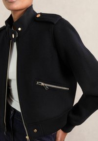 Veste à zip en laine noire avec des boutons-pression dorés et une poche zippée, portée sur une chemise blanche et un jean foncé, coupée au milieu du torse.