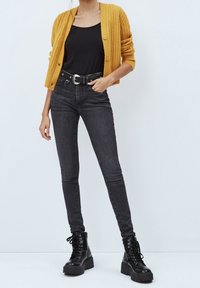 Person som bär en senapsgul cardigan över en svart topp, högmidjade mörkgrå tajta jeans med bälte, och svarta snörade platåstövlar.