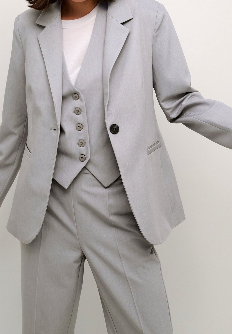 Costume gris clair comprenant un blazer ajusté, un gilet à cinq boutons et un pantalon à jambes larges. Tissu lisse avec une texture subtile et des poches.