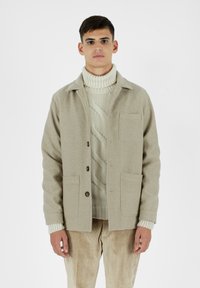 Giacca di lana beige con colletto, chiusura a bottoni, due tasche frontali, indossata sopra un dolcevita in maglia crema, abbinata a pantaloni in velluto a coste.