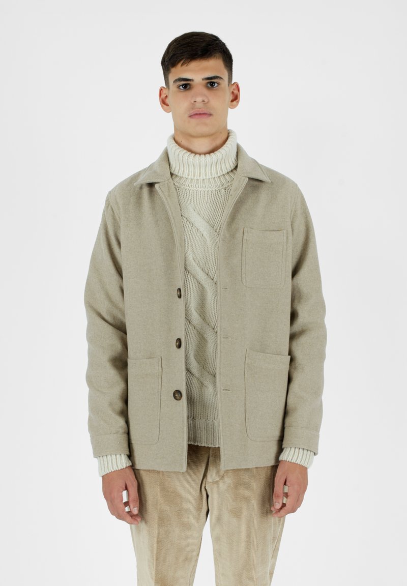 Giacca di lana beige con colletto, chiusura a bottoni, due tasche frontali, indossata sopra un dolcevita in maglia crema, abbinata a pantaloni in velluto a coste.