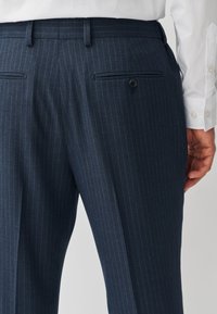 Uomo che indossa pantaloni eleganti blu navy a righe sottili con tasca posteriore con bottone e camicia bianca, mostrato dalla vita a metà coscia.