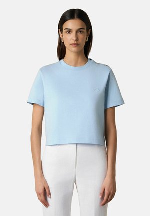 Jeune femme aux cheveux raides et foncés, portant un haut bleu clair à manches courtes avec des détails de boutons sur l'épaule et un pantalon blanc.