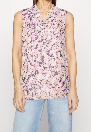 Ärmellose Bluse mit abstraktem Blumenmuster in Rosa, Lila und Beige, kombiniert mit hellblauen Jeans an einer stehenden Frau.