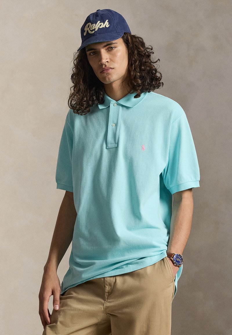 Mann mit langen lockigen Haaren, trägt eine blaue Ralph-Kappe, ein hellblaues Poloshirt, beige Hose und eine blaue Armbanduhr, steht vor einem neutralen Hintergrund.